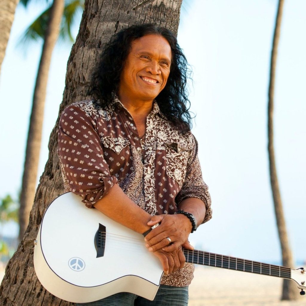 An Image of Henry Kapano at the beach, holding a white guitar, while standing under a Palm Tree.