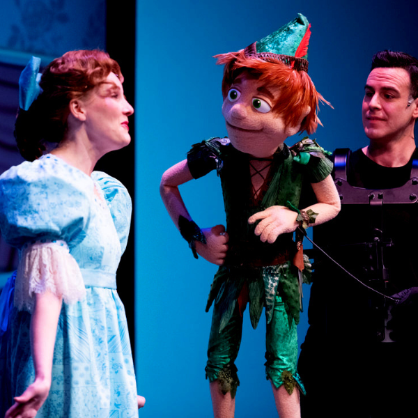 DLUX Puppets Neverland 600x600 Main Image. Image of two people: a woman in a blue dress and a man in black holding a Peter Pan Puppet.