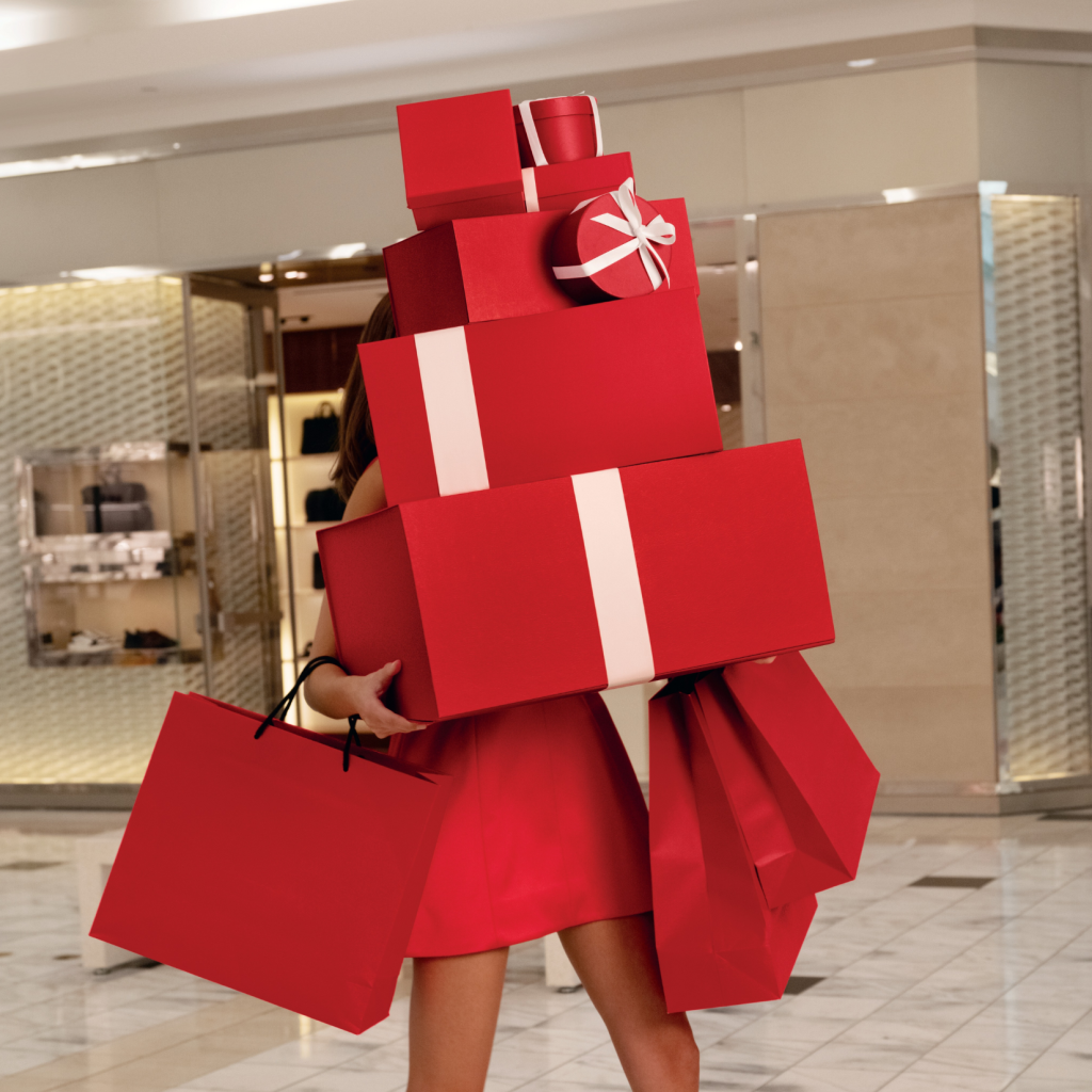Woman in red dress with red packages and white bows stacked high covering her face walking through mall.