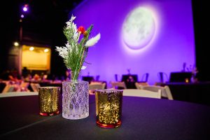 A vase with dried flower and two candles on a table with a moon on purple background.